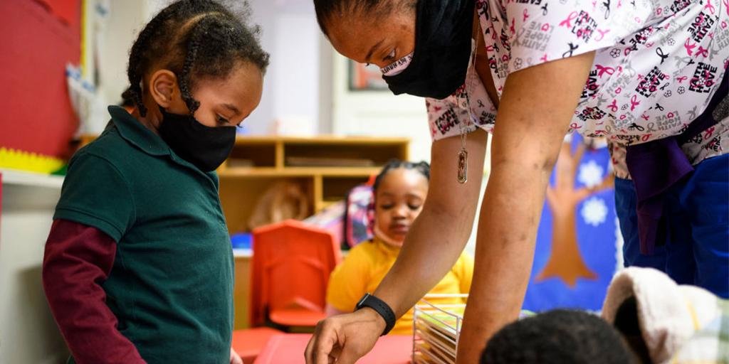 A teacher wearing a mask helps a young student